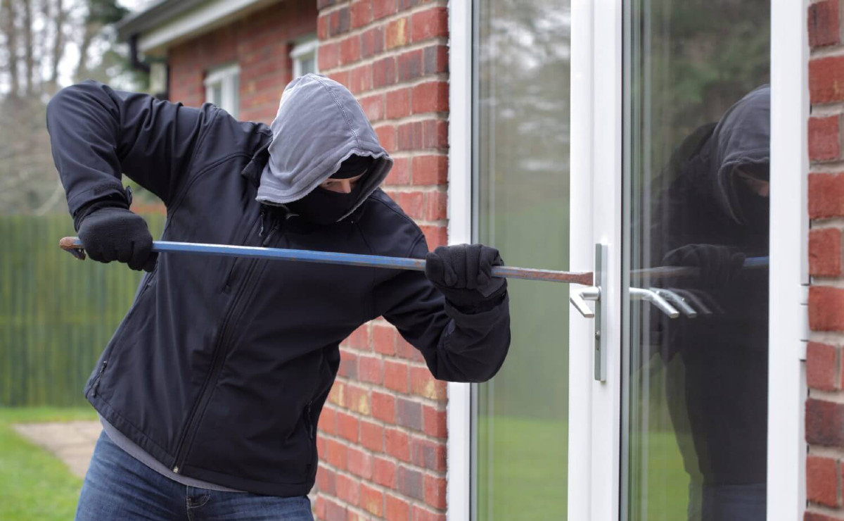 Person in black sweatshirt and gray hoodie uses crowbar to try and open a patio door of a red-brick home.
